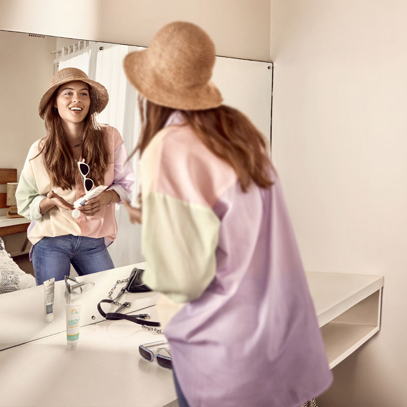 A young woman smiling at her reflection in her bathroom mirror while she gets prepared to heard outdoors with her hat, sunglasse and sunscreen